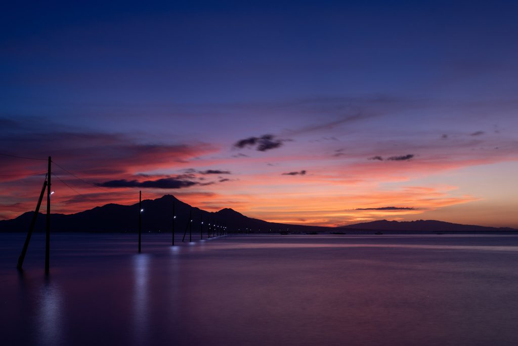 夕焼けに染まる有明海と、明かりが灯る長部田海床路の電柱、遠くに連なる山のシルエット