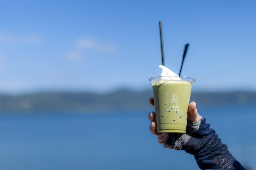 池田湖を背景に手に持った、ソフトクリームをのせた抹茶ドリンク