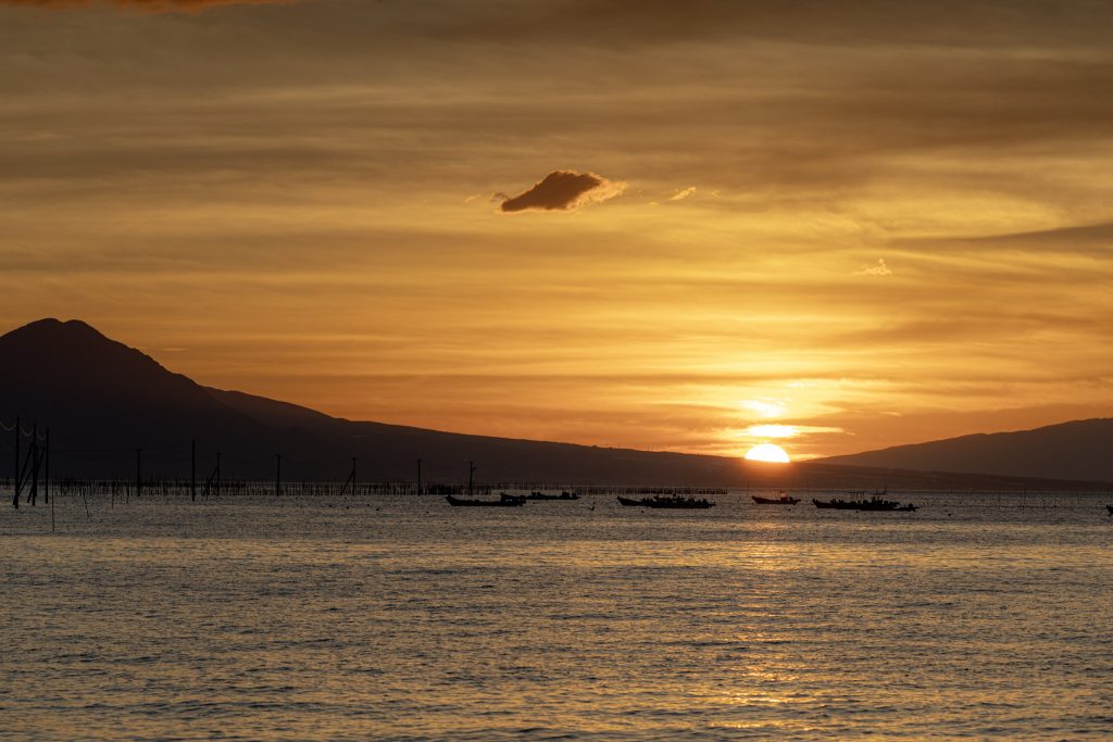 夕日が山の間へ沈む有明海と、水面に浮かぶ船のシルエット
